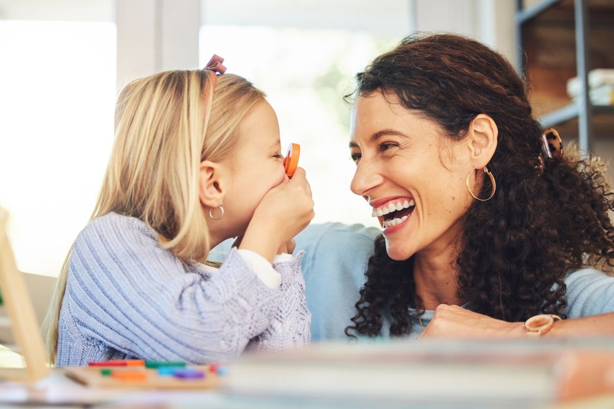 Little girl whispering a secret or joke into her mother's ear while her mother laughs at home.