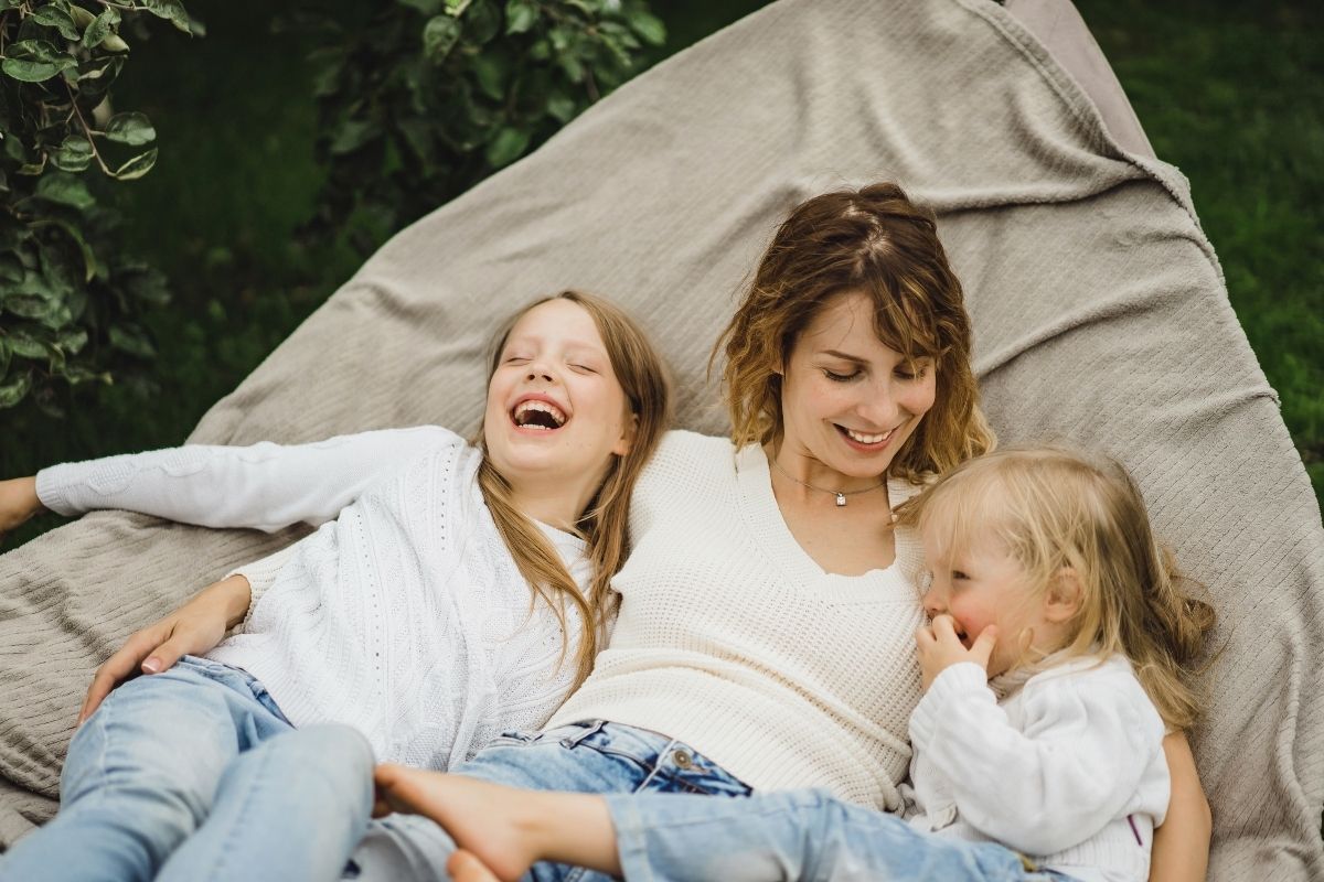 Mother relaxing outdoors on a large cushion with her two daughters, one laughing and the other smiling beside her.
