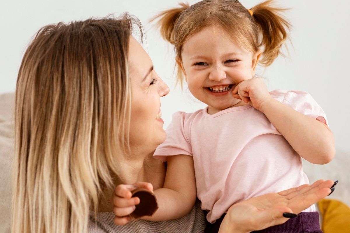 Smiling mother holding her laughing young daughter in a pink shirt, sharing a playful moment together at home.