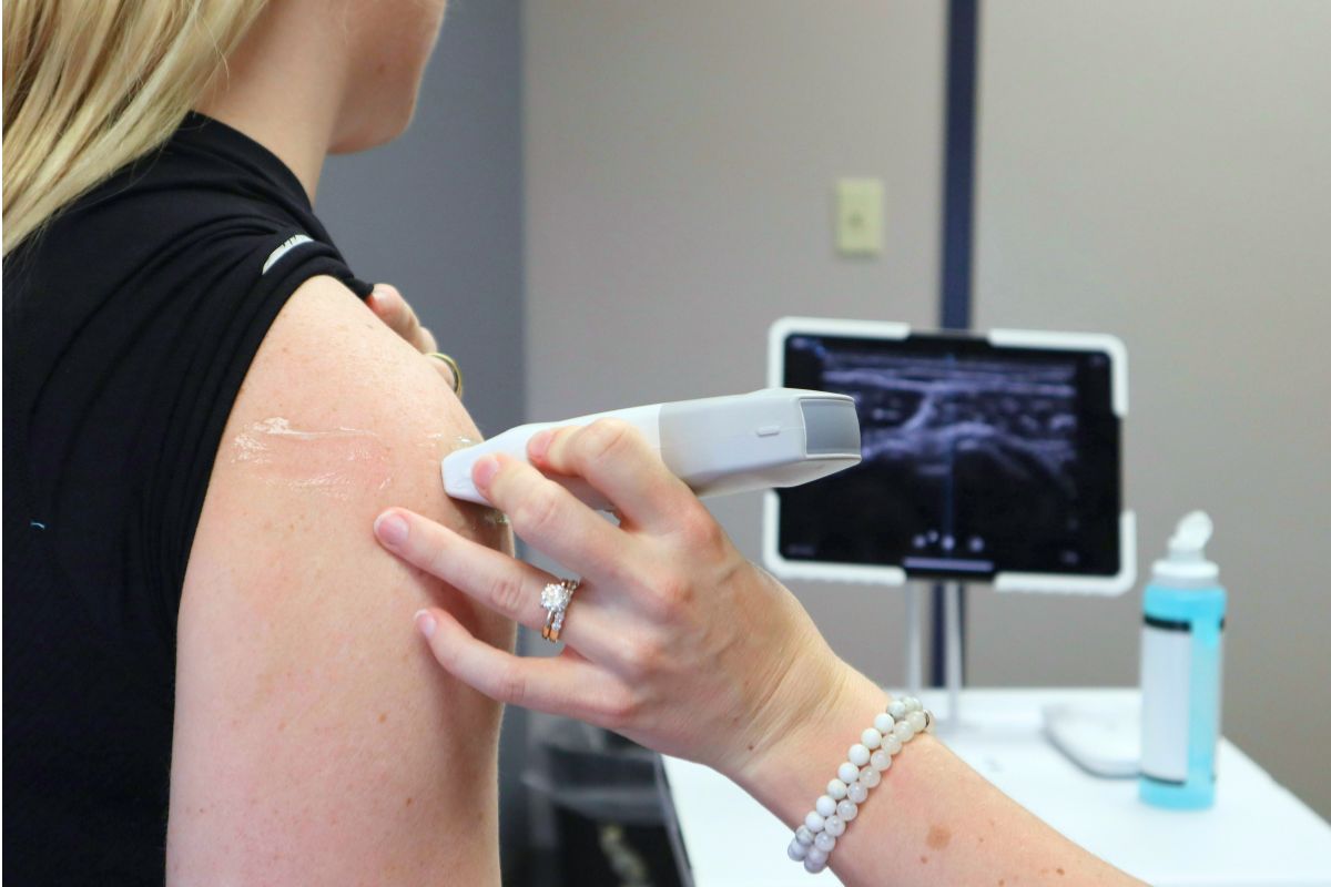 Healthcare provider using an ultrasound device on a patient’s shoulder in a clinical setting.