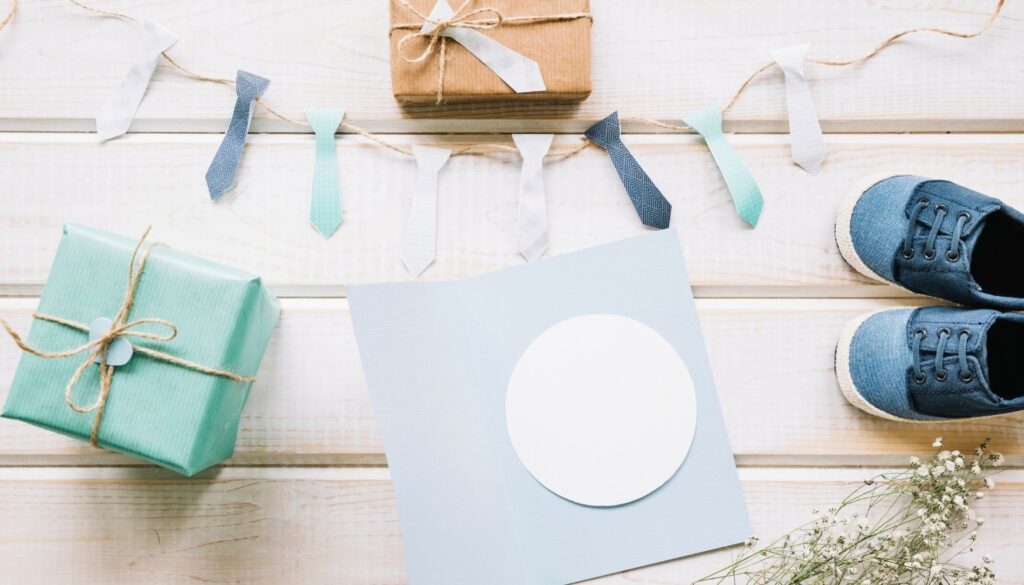 Flat lay of baby shower gifts, a blank greeting card, baby shoes, and decorative paper ties on a light wooden background.