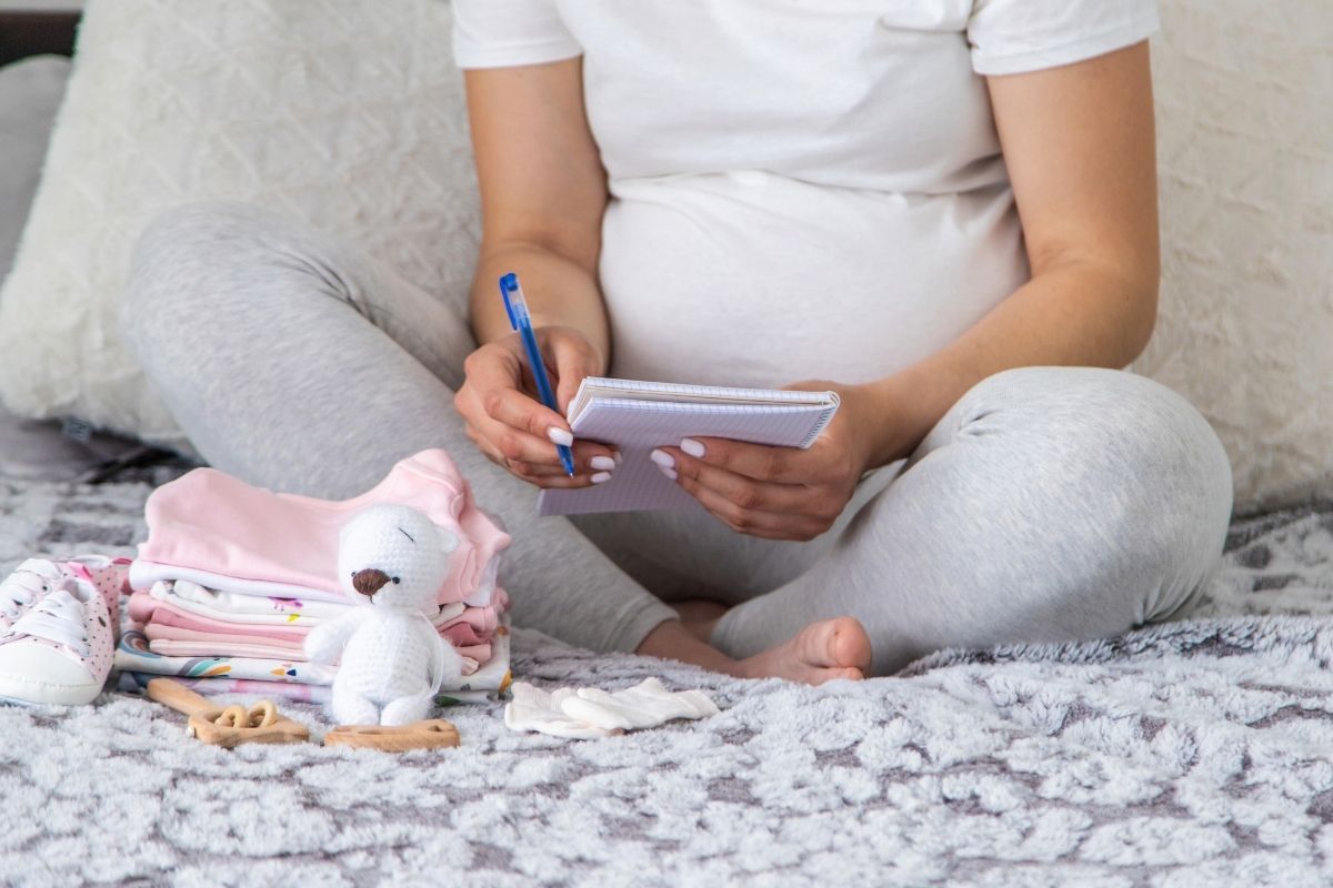 Pregnant woman writing a baby registry list beside folded baby clothes and a small teddy bear.