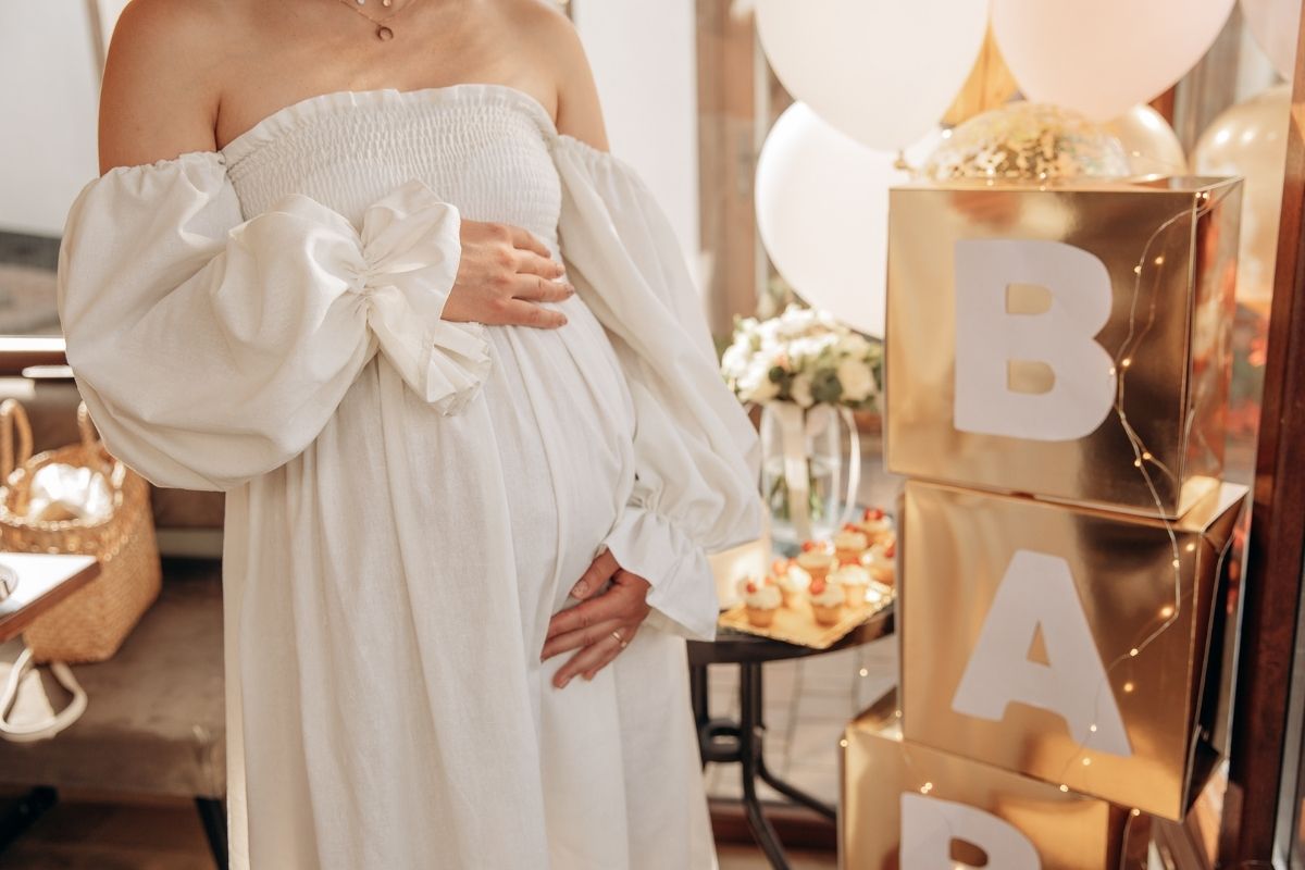Pregnant woman wearing a white off-the-shoulder dress at a baby shower celebration, surrounded by balloons and decorative baby blocks.