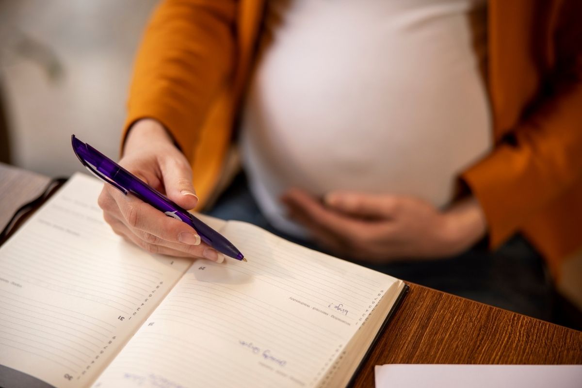 Pregnant woman in a mustard cardigan writing in an open planner with a purple pen while resting her other hand on her belly.