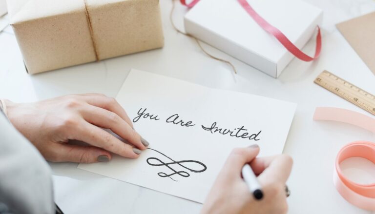 Close-up of a person handwriting a "You Are Invited" card at a desk with wrapped gifts, ribbons, and stationery supplies.