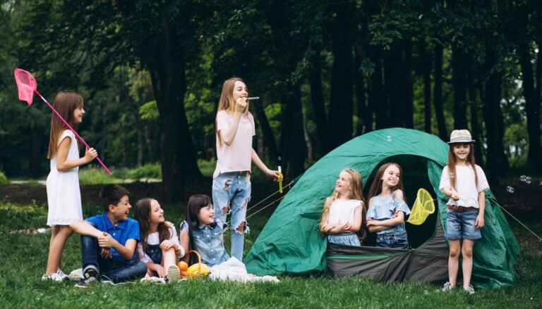 A group of children enjoying outdoor camping activities with butterfly nets and bubbles beside a green tent in a wooded campsite.
