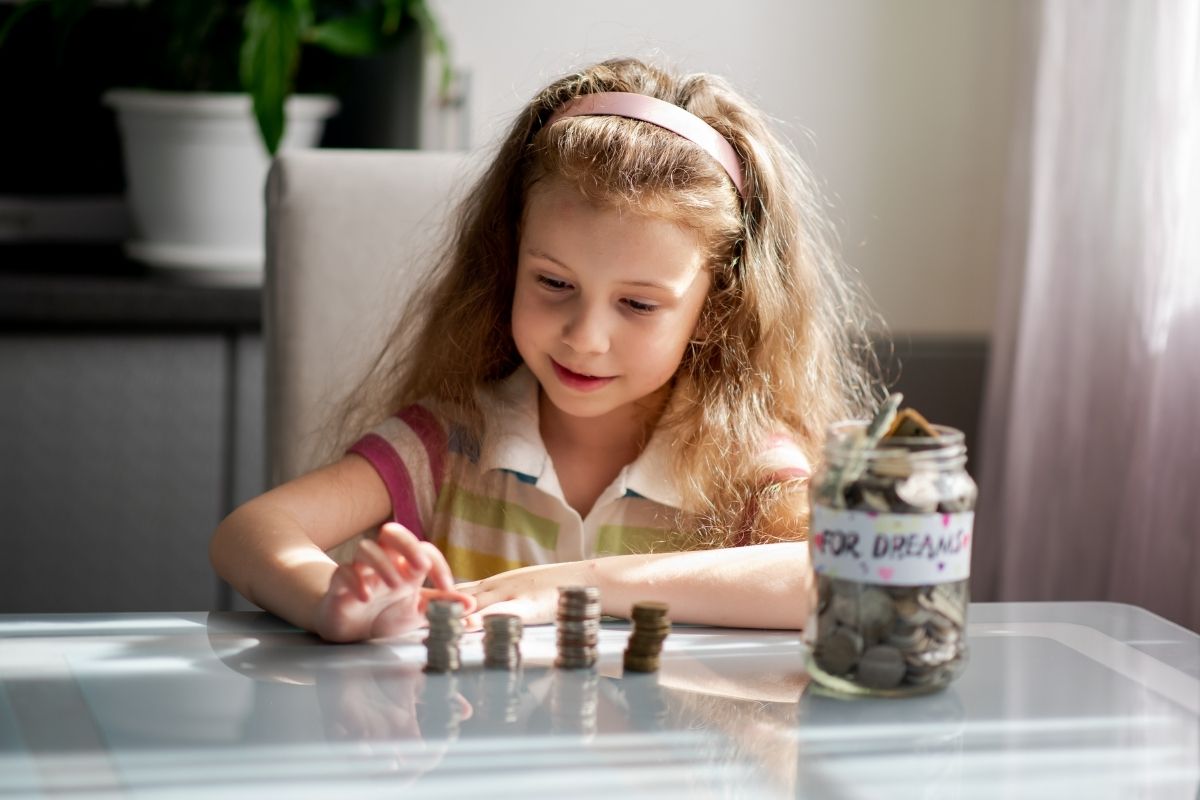 Young girl stacking coins beside a savings jar at a table.