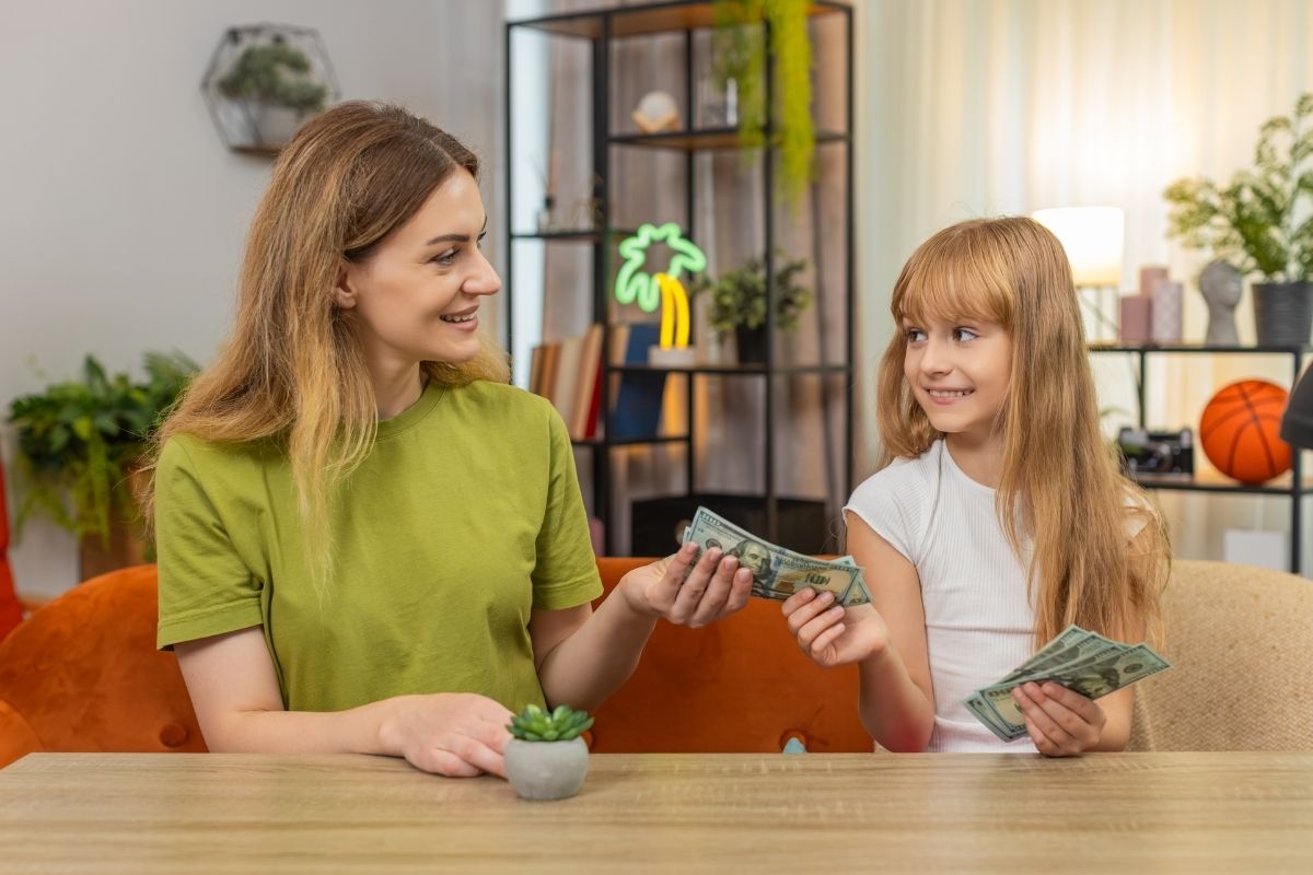 Mother giving cash to her daughter while sitting together at a table.