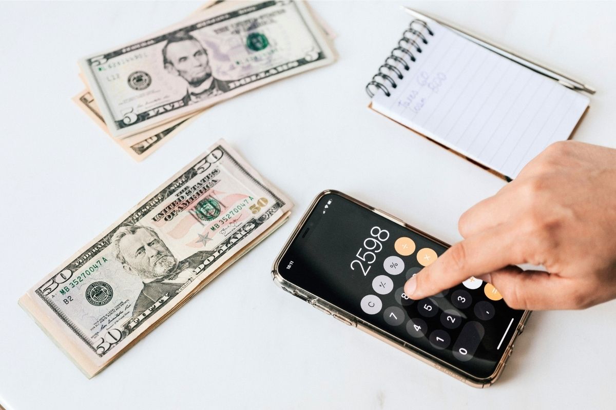Hand using a calculator next to dollar bills and a notebook.