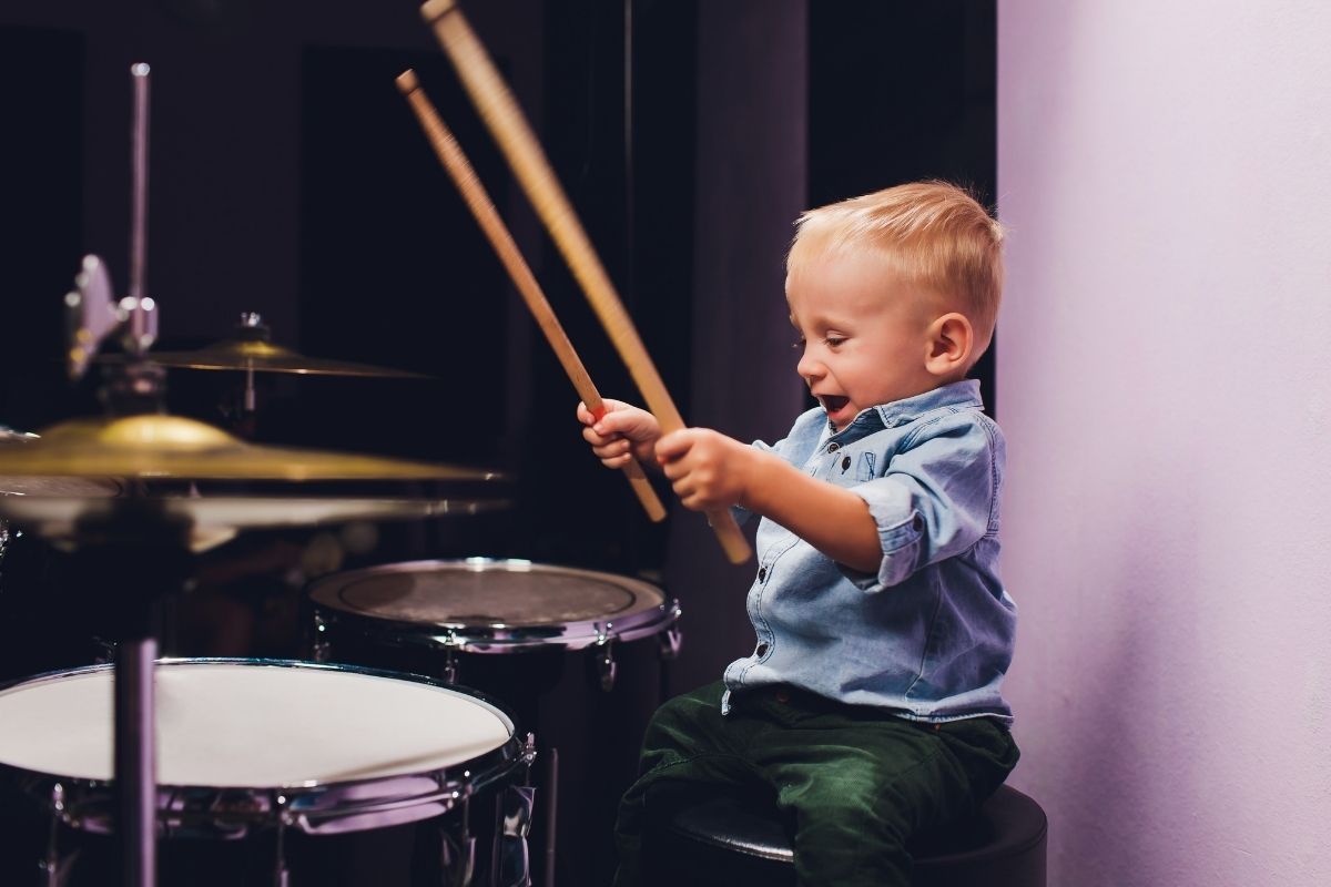 Young boy smiling while playing a drum set with drumsticks.