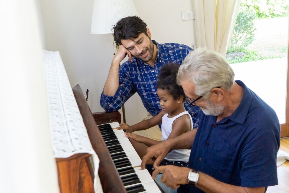 Grandfather and granddaughter playing the piano together while her father watches.