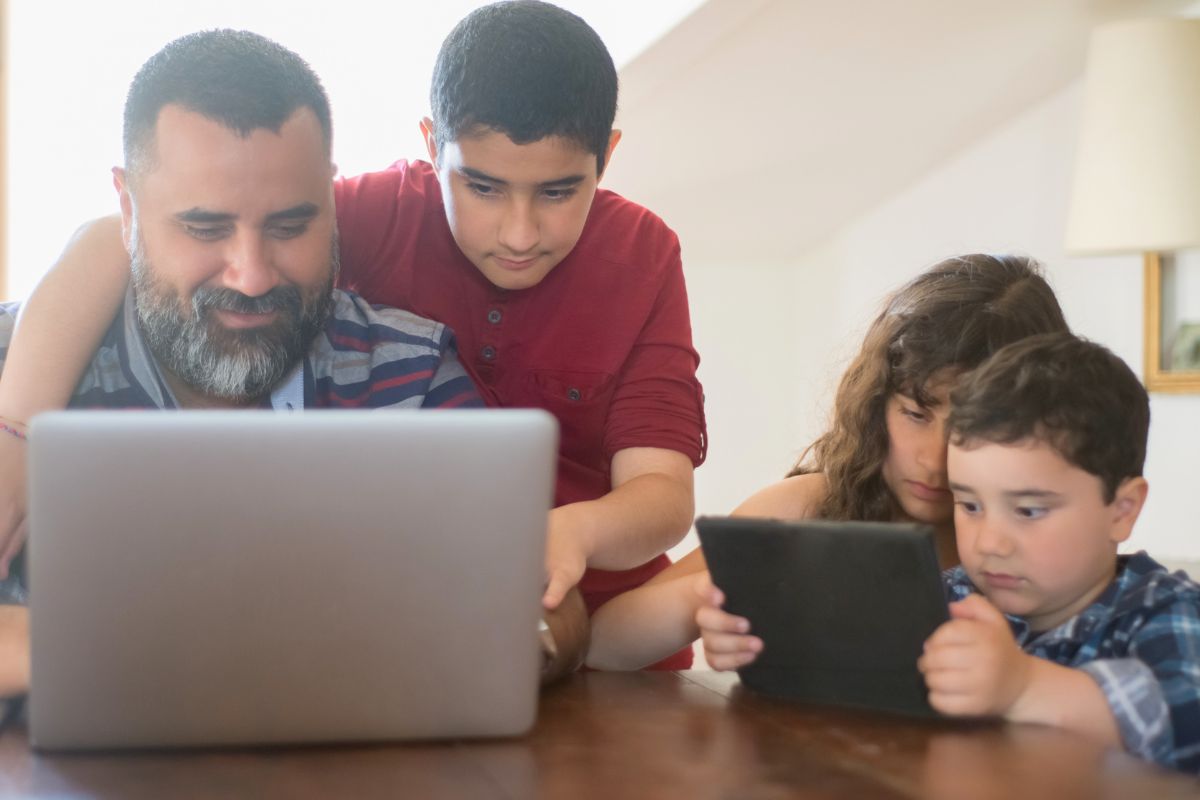 Father supervising children using a laptop and tablet at home.