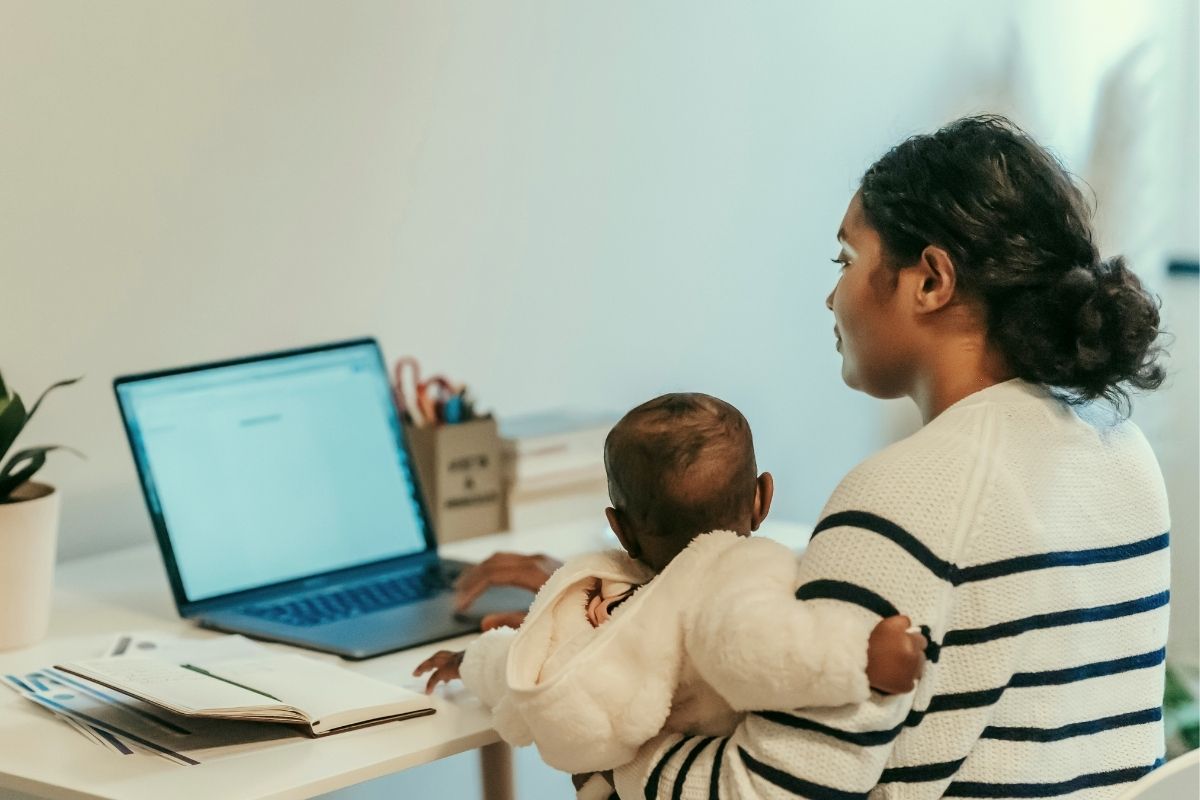 Working parent holding a baby while using a laptop at home office setup.