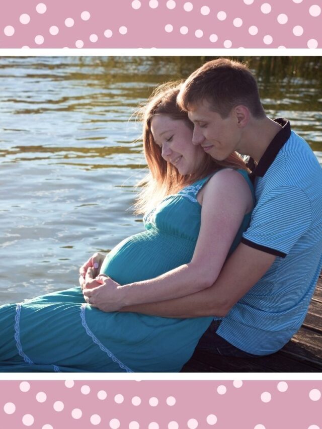Pregnant couple sitting together by the water, with the partner hugging the mother’s baby bump.