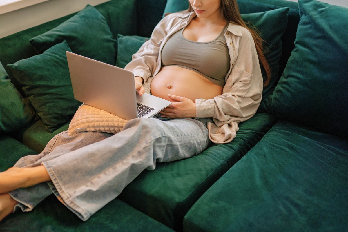 Pregnant woman resting on a green couch while using a laptop during babymoon planning.