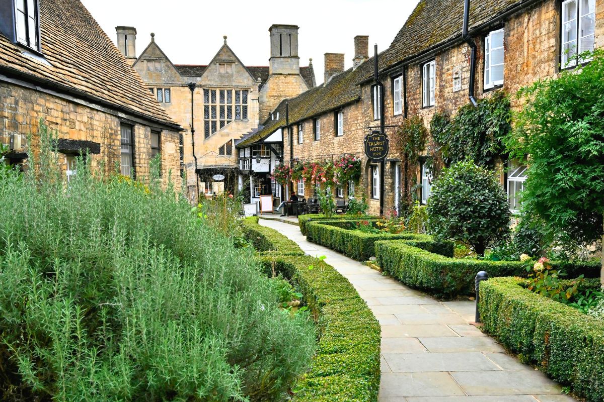 Peaceful Cotswolds village walkway with stone buildings, greenery, and cottage-style charm.