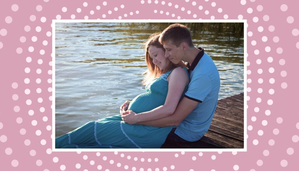 Pregnant couple sitting together by the water, with the partner hugging the mother’s baby bump.