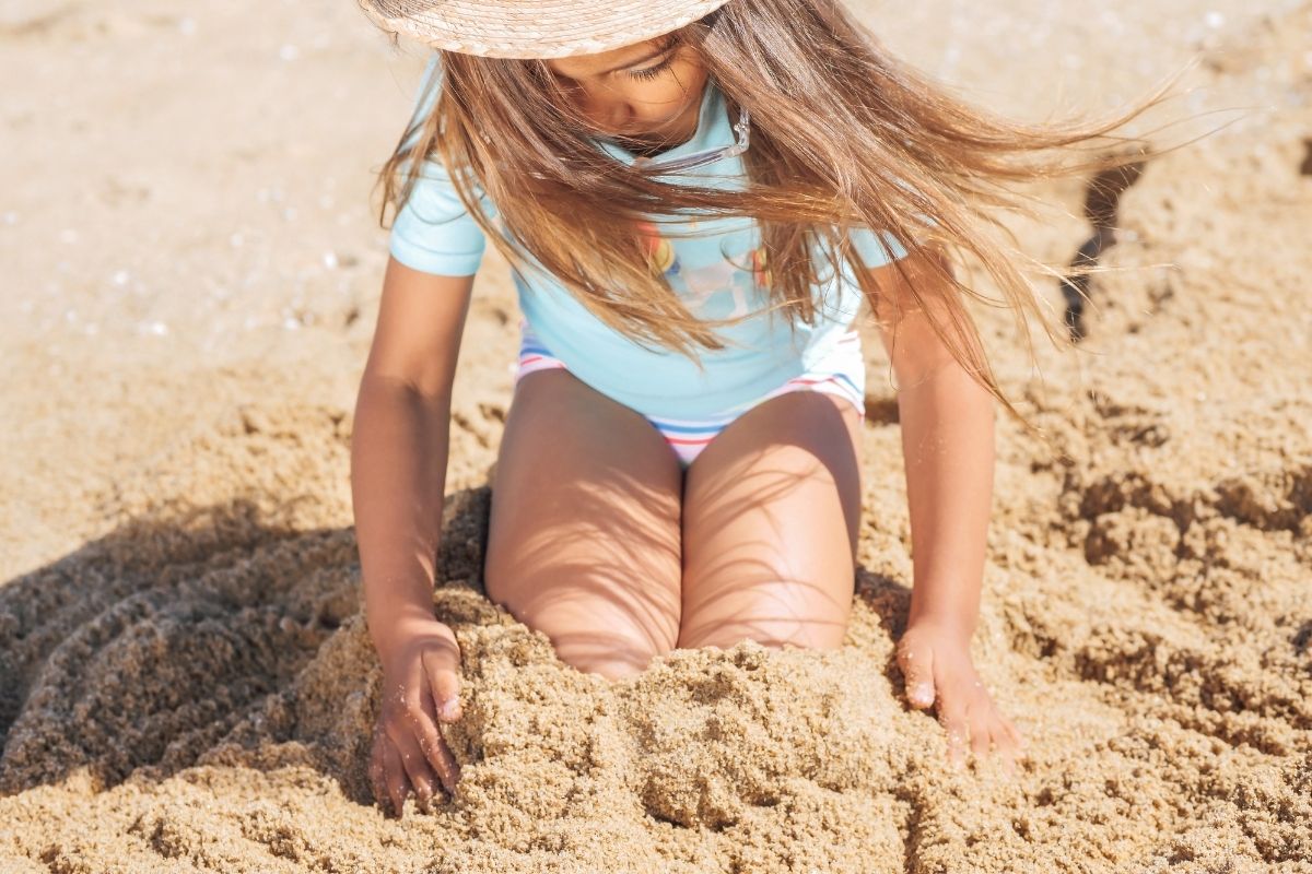 Toddler playing in sand at the beach wearing sun hat, highlighting essentials for toddler beach vacation packing list.