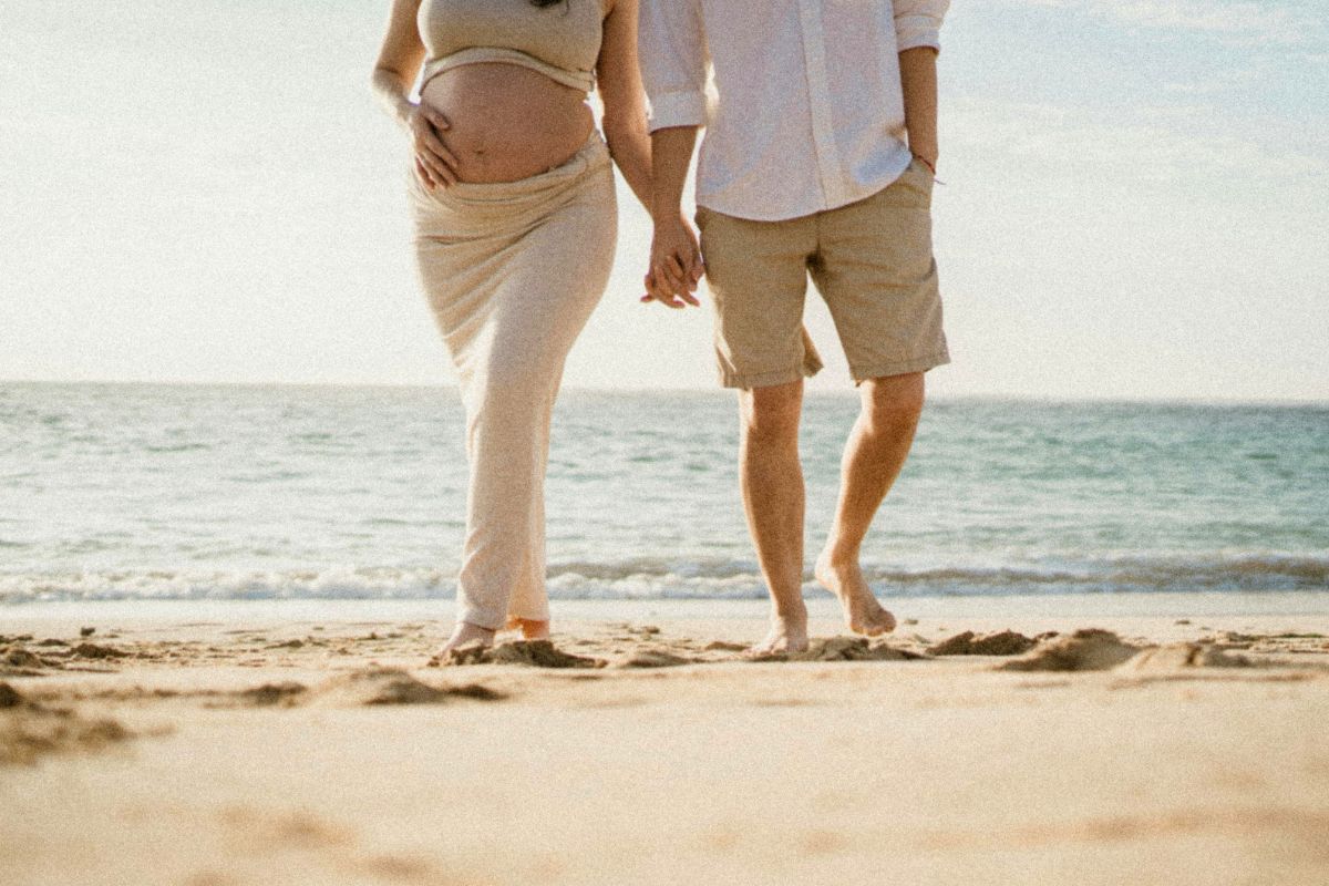 Pregnant couple holding hands while walking on the beach, enjoying a peaceful babymoon date before baby arrives.