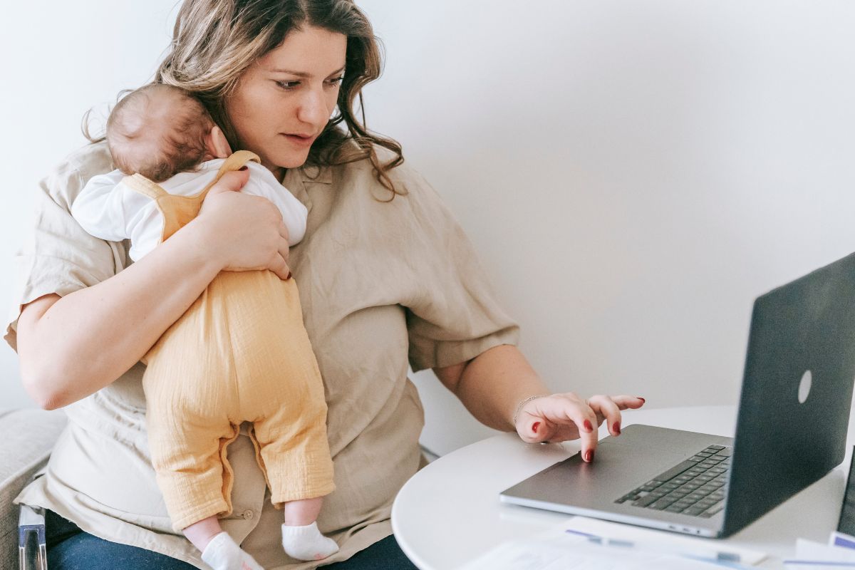 New mother holding her baby while using a laptop to prepare for returning to work.