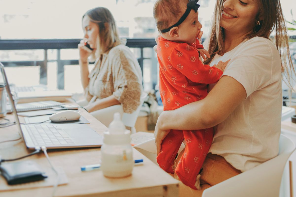 Working mother holding her baby beside a laptop, bottles, and other feeding supplies.