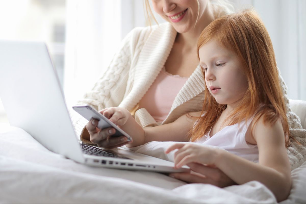 Mother teaching her young daughter how to use AI safely on a laptop and smartphone at home.