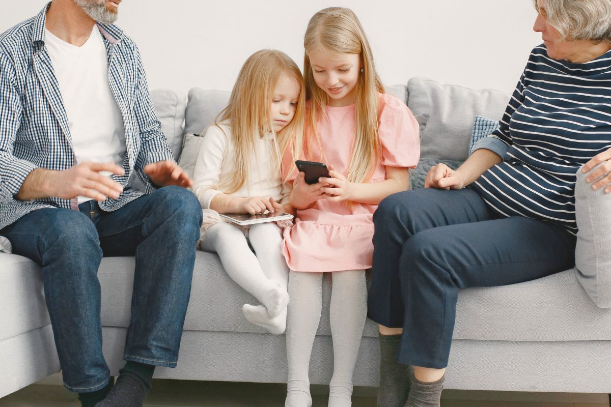 Parents supervising two young girls as they use a smartphone and tablet on the couch.