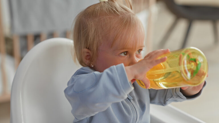 Toddler drinking from a large water bottle while sitting in a high chair.