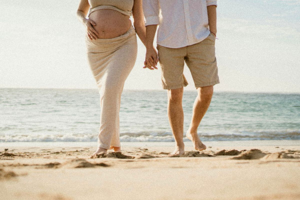 Expecting couple walking hand in hand on the beach during a relaxing babymoon getaway.