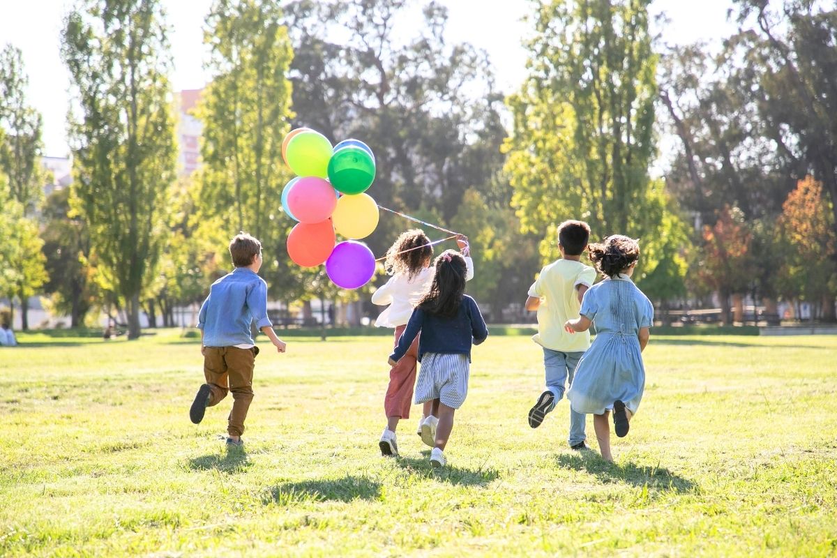 Group of children running across a sunny grassy field with balloons.