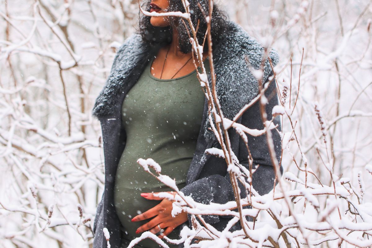 Pregnant woman in a warm coat standing among snowy trees during a cozy winter babymoon.