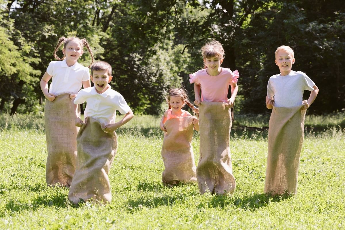 Five children enjoying a sack race outdoors on a sunny day, jumping across a grassy field.
