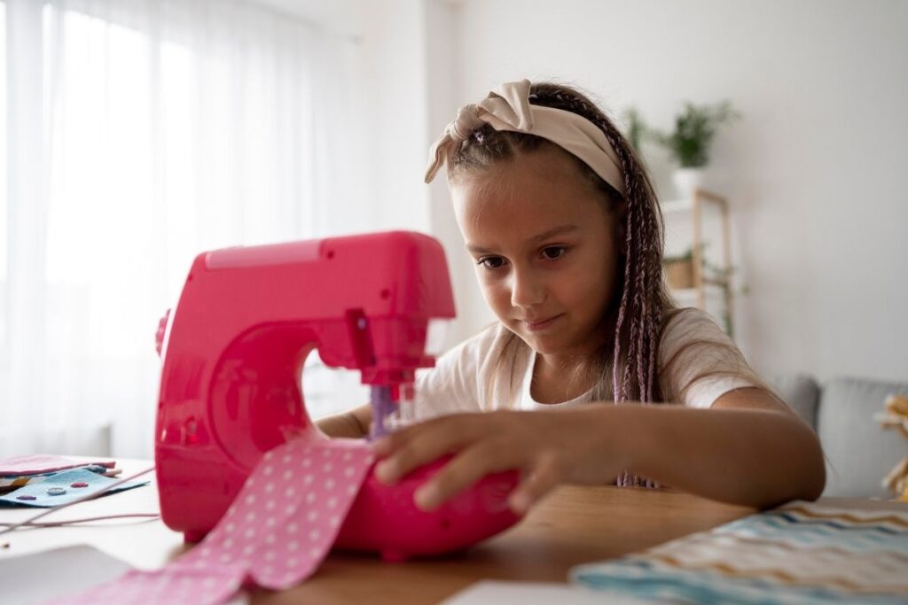 Young girl using a pink sewing machine to sew polka-dot fabric at home.