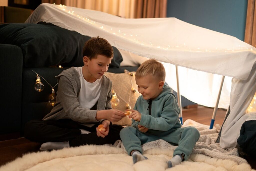 Two young boys playing together inside a homemade blanket fort decorated with warm string lights.
