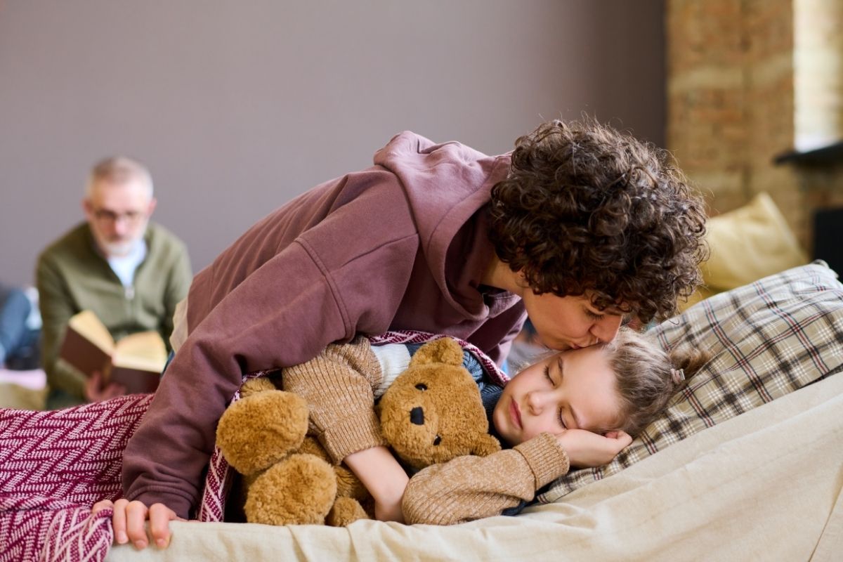 Parent gently kissing a sleeping child on the forehead while the child cuddles a teddy bear in bed.