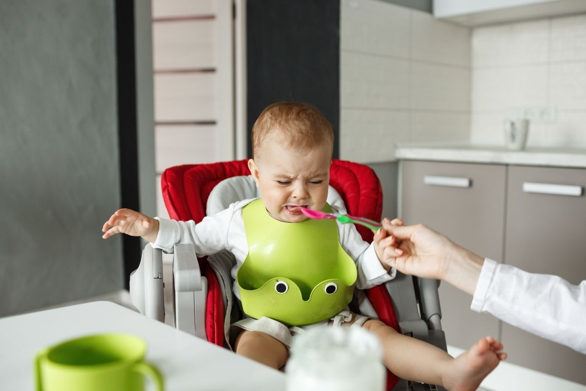 Baby sitting in a high chair wearing a green bib, turning away from a spoon offered by an adult during feeding.