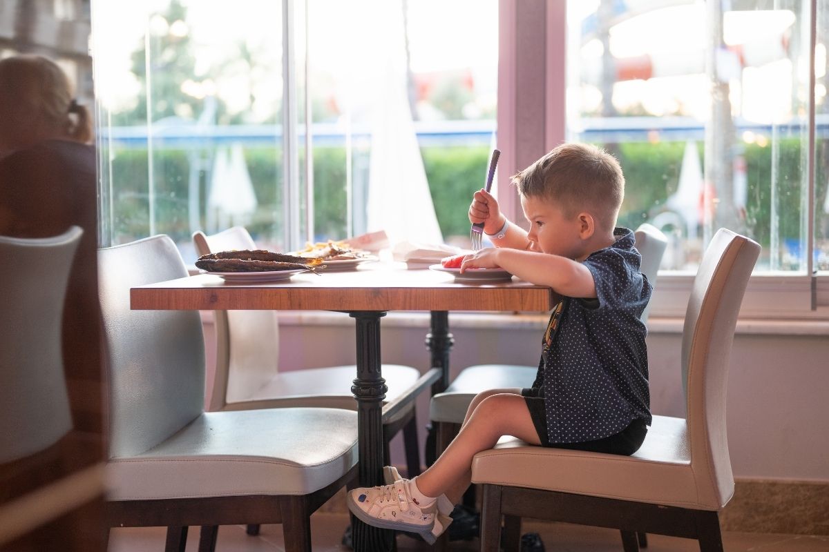 Young boy sitting at a restaurant table by a large window, leaning forward while eating with a spoon.