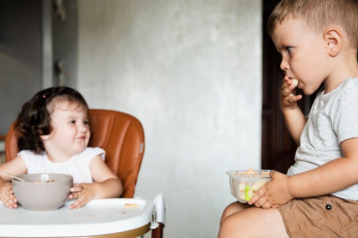 Two young children eating together, with a toddler in a high chair watching an older boy sitting nearby with a snack.
