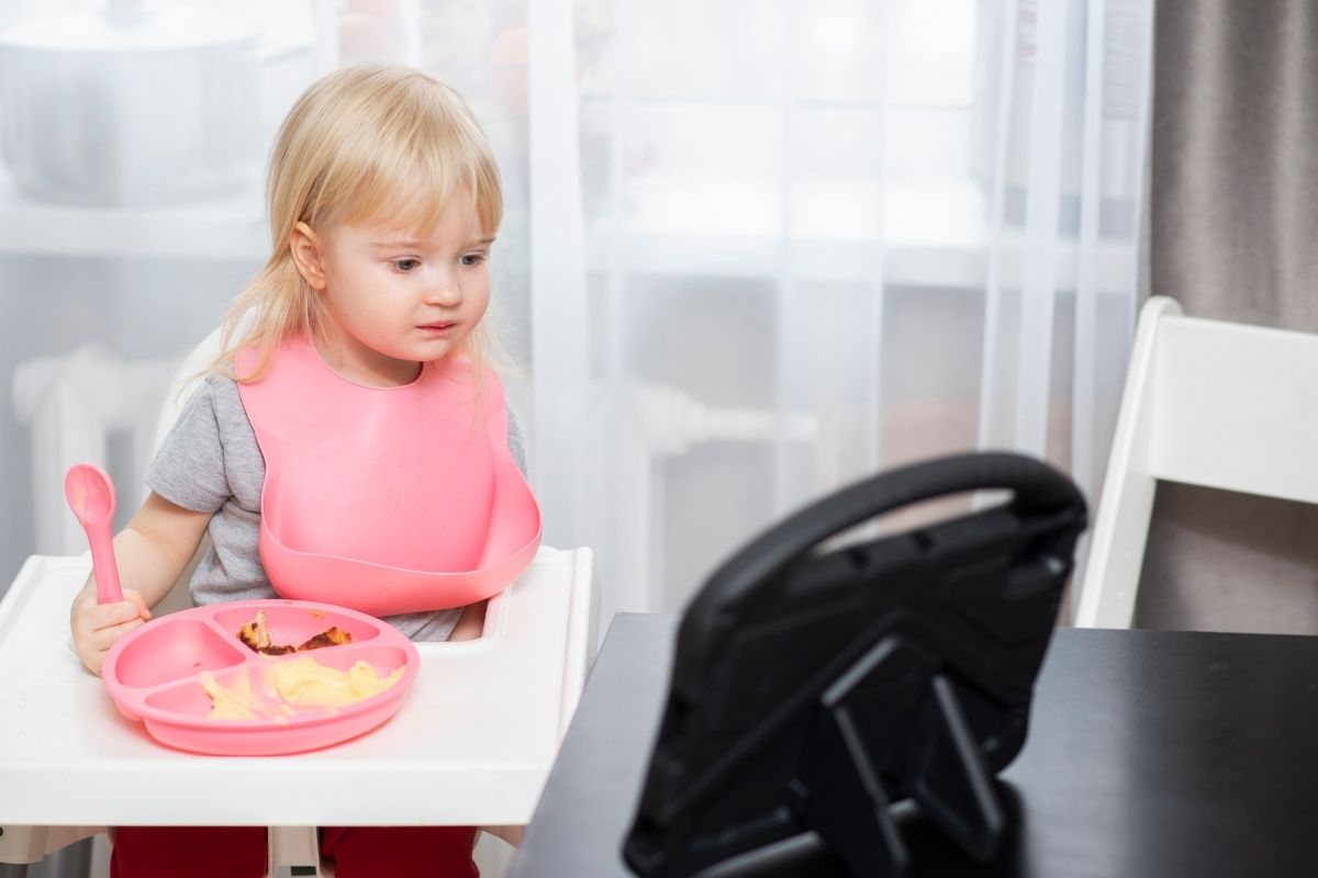 Little girl sitting in a high chair with a pink plate and spoon, looking at a tablet during mealtime.