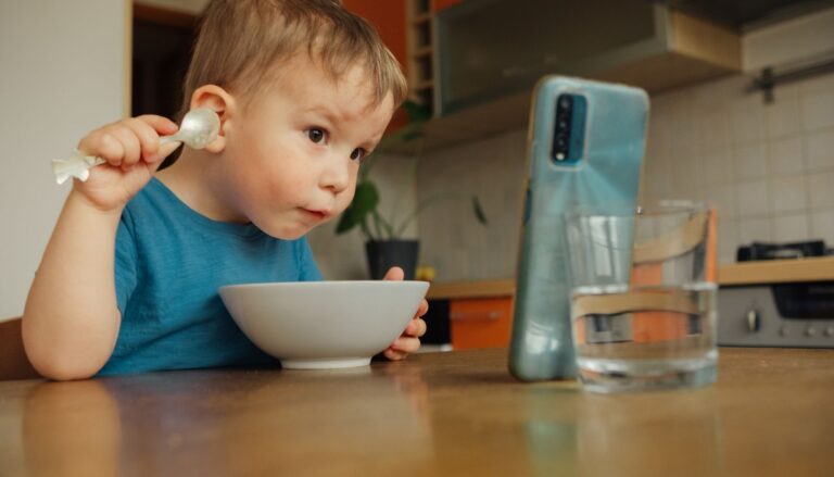 Toddler sitting at a kitchen table with a spoon and bowl, distracted by a smartphone propped up in front of him during mealtime.