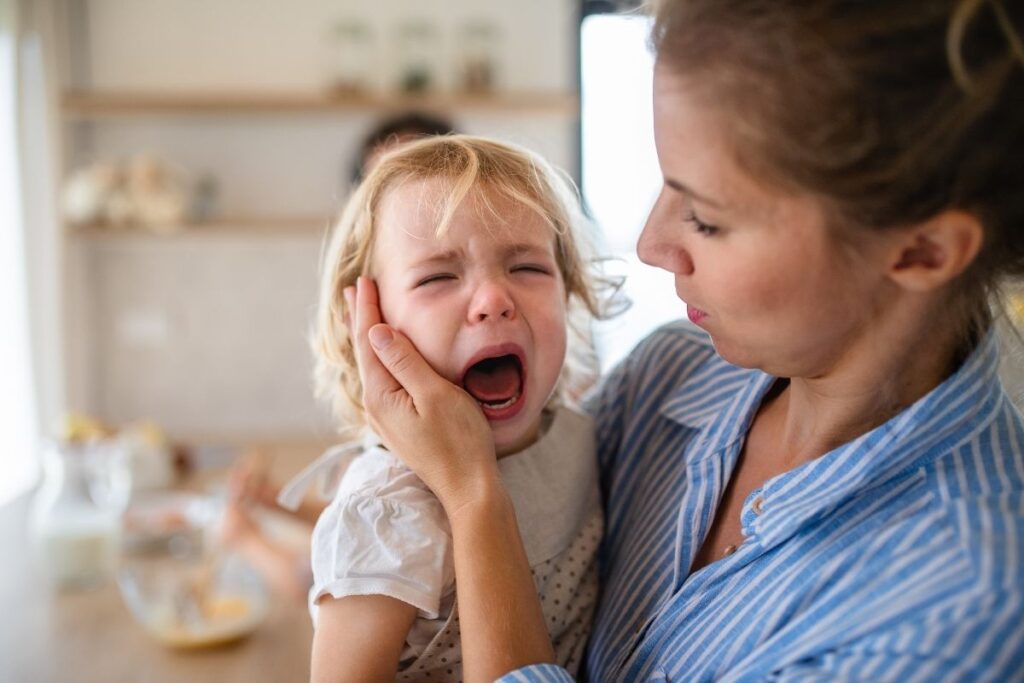 Mother gently comforting her crying toddler by holding the child's cheek during an emotional moment at home.