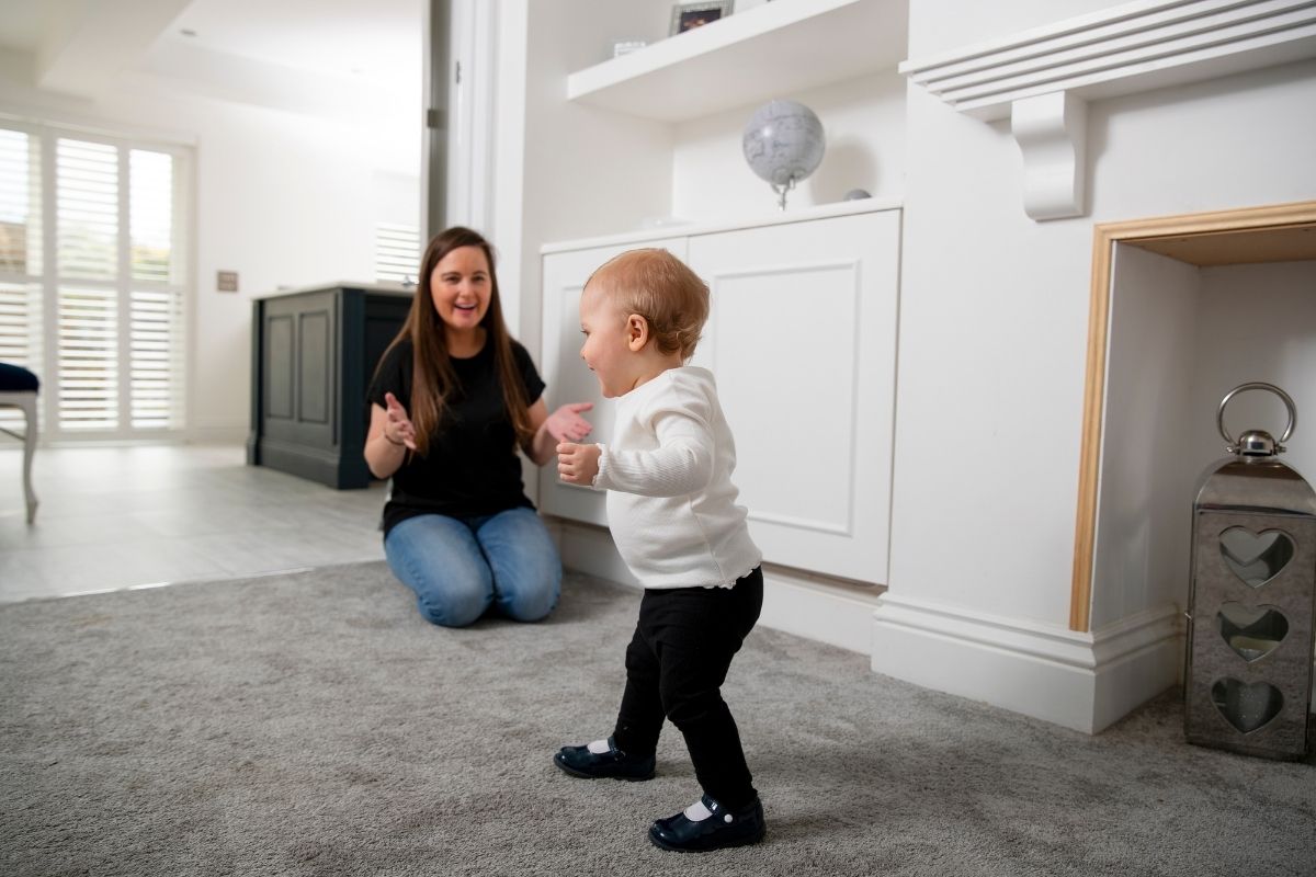 Toddler taking steps across a living room toward his smiling mother, who is kneeling nearby with her arms open.