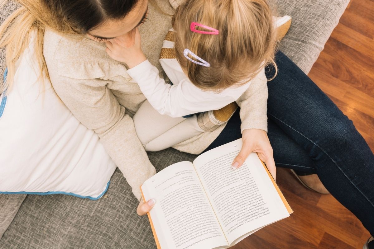 Overhead view of a mother sitting with her young daughter on a sofa, sharing a quiet moment while holding an open book.