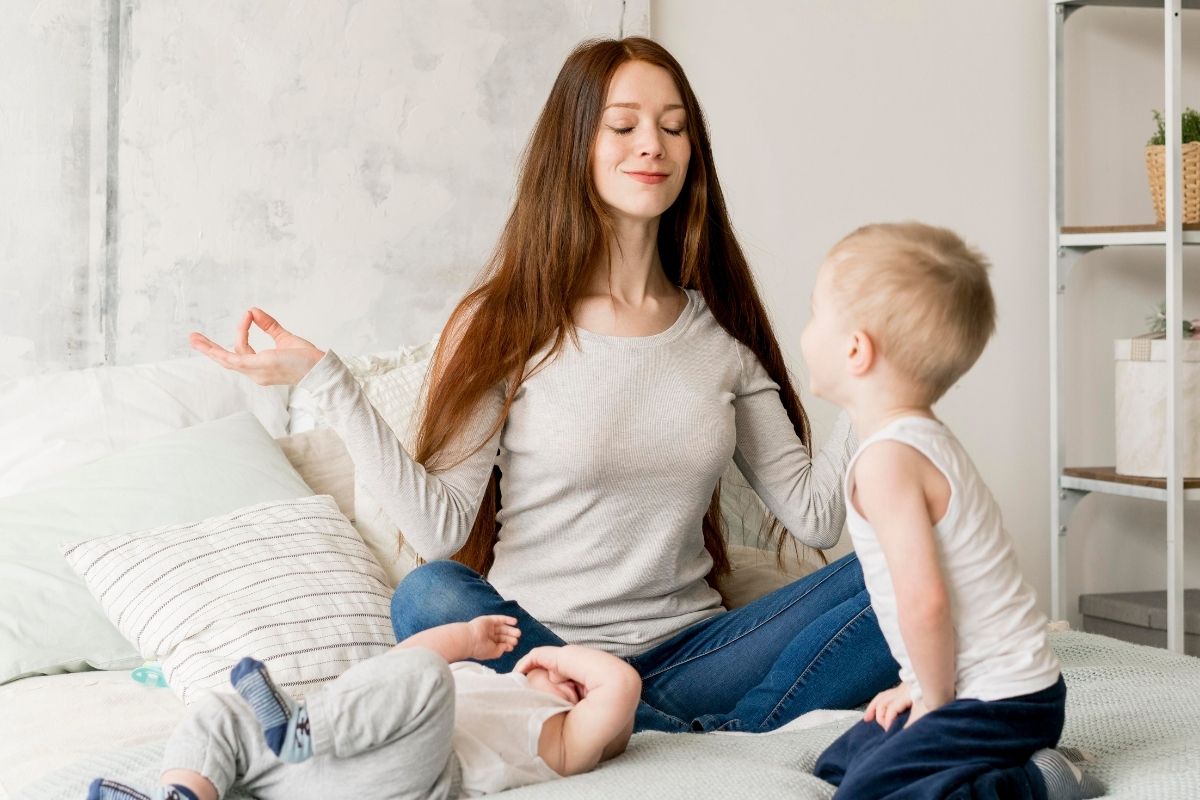 Mother sitting cross-legged on a bed with her eyes closed and hands in a meditation pose while two young children play beside her.
