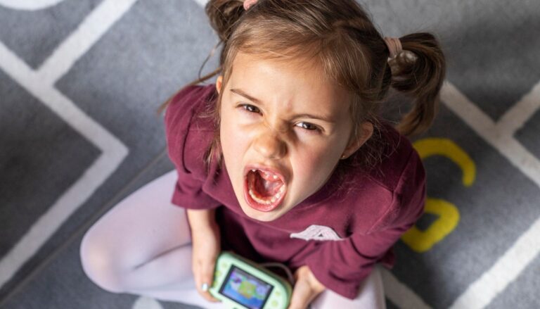 Young girl sitting on the floor, screaming with frustration while holding a smartphone, illustrating a toddler tantrum.