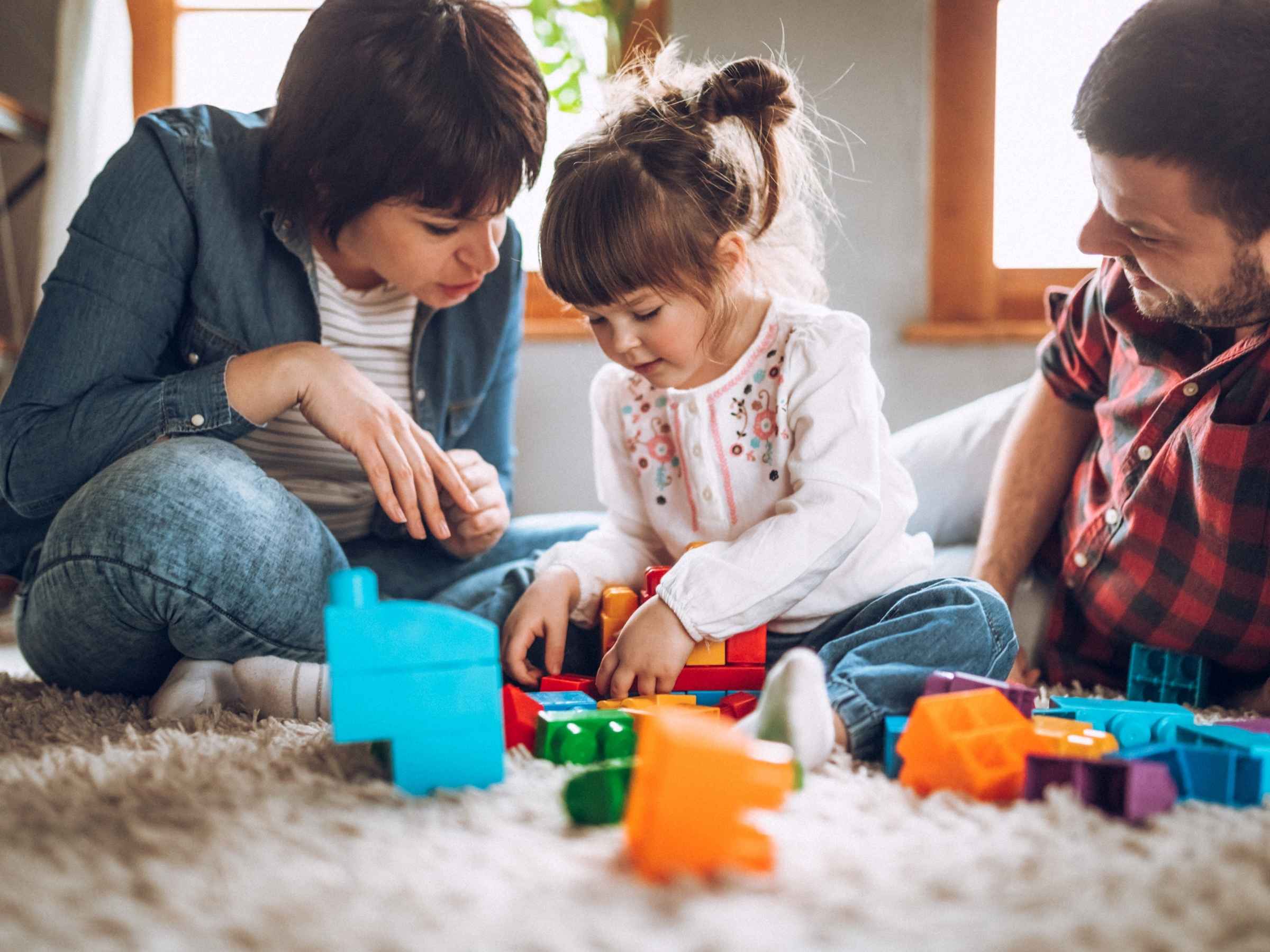 Mother, father, and toddler sitting on a carpeted floor, playing with colorful plastic building blocks