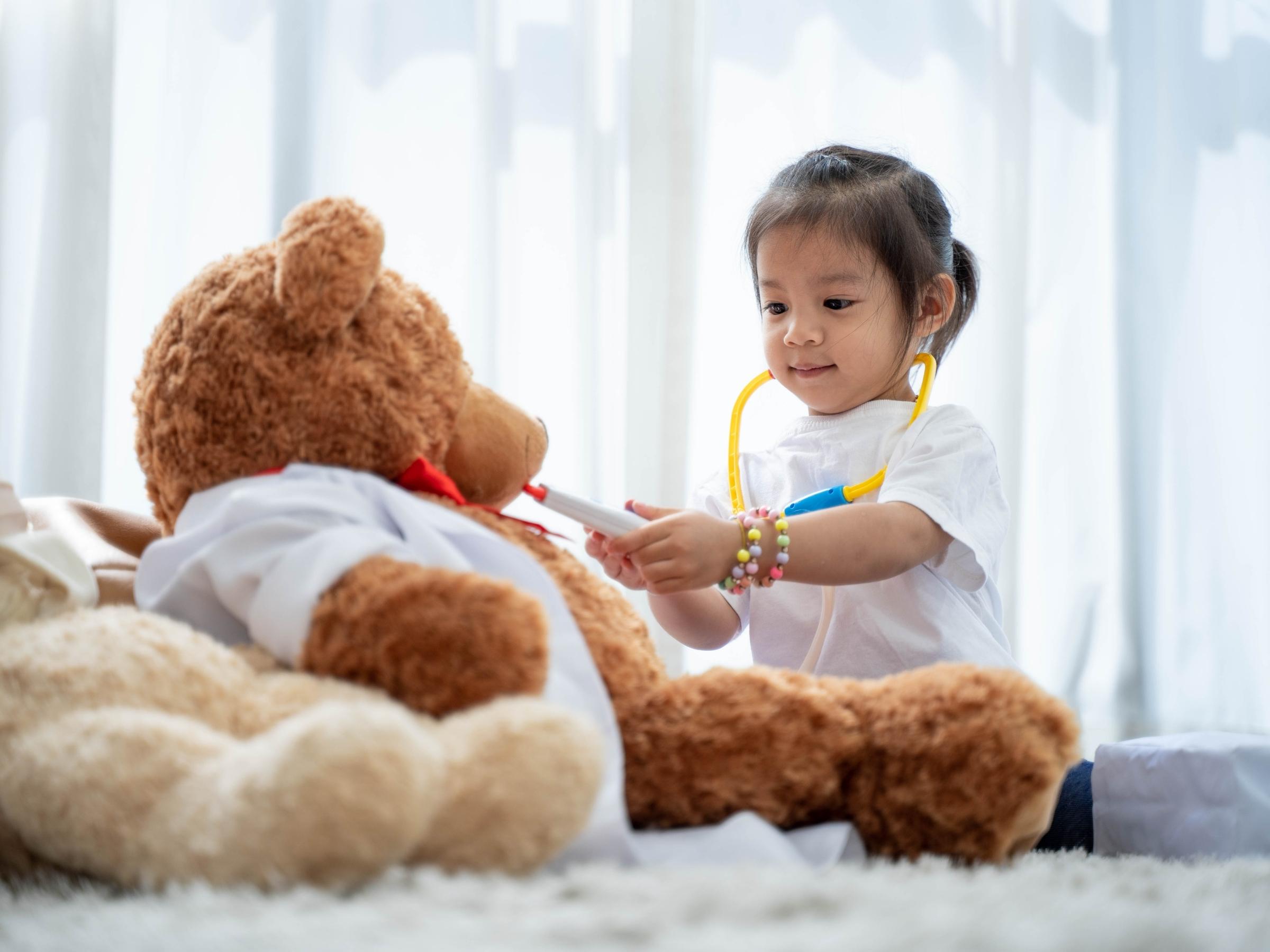 a girl toddler engaged in imaginative play, pretending to give a teddy bear a medical check-up using a toy stethoscope.