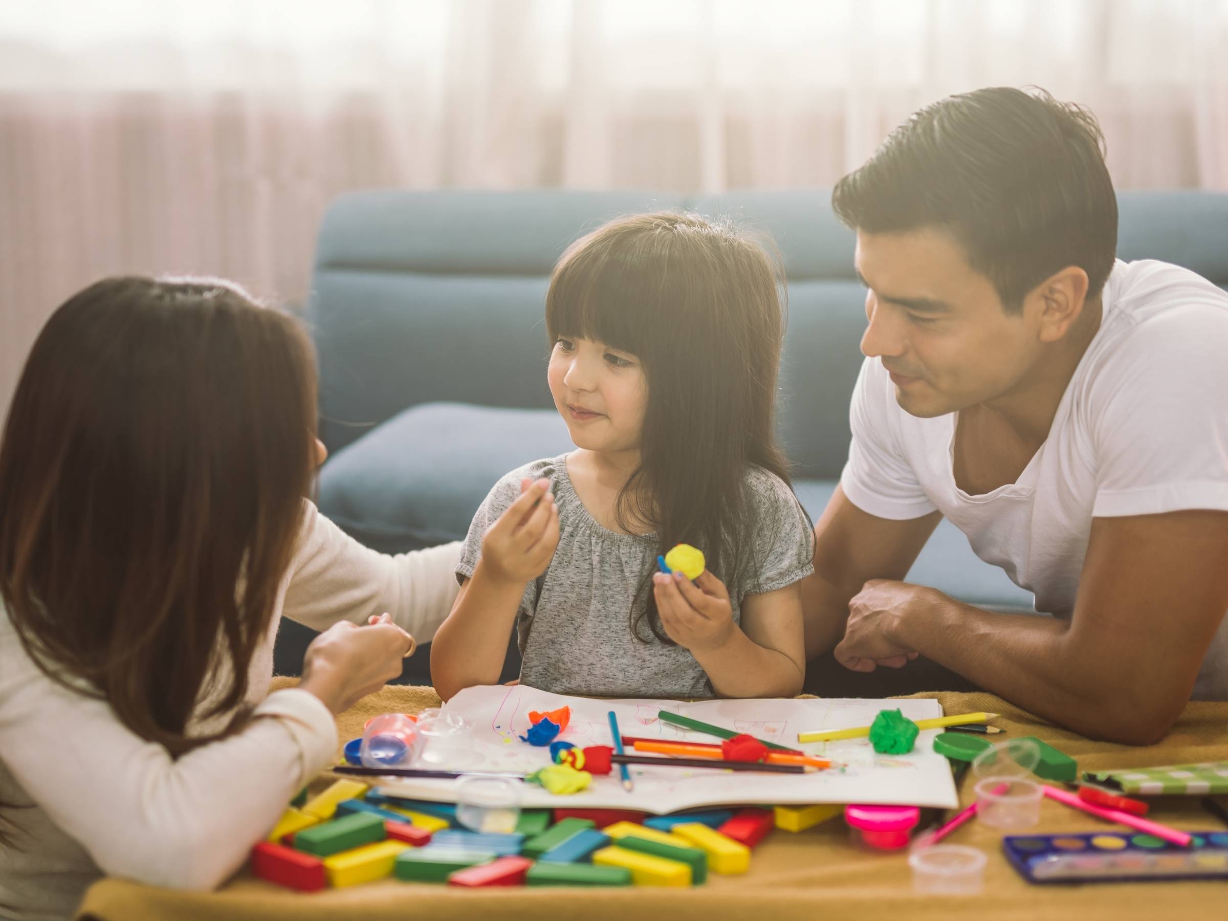 Mother and father sitting at a table with their toddler daughter in the middle, engaged in a creative activity with colorful modeling clay, paintbrushes, markers, and building blocks