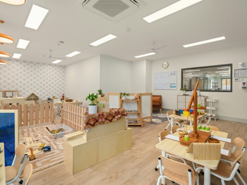 Children's room equipped with small tables and chairs, educational toys, a sandbox play area, and decorative plants