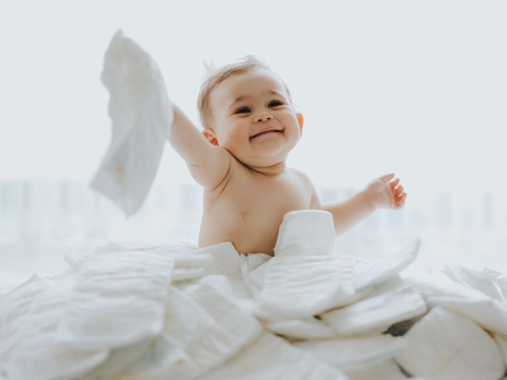 Baby sitting among a large pile of diapers, holding one up in the air